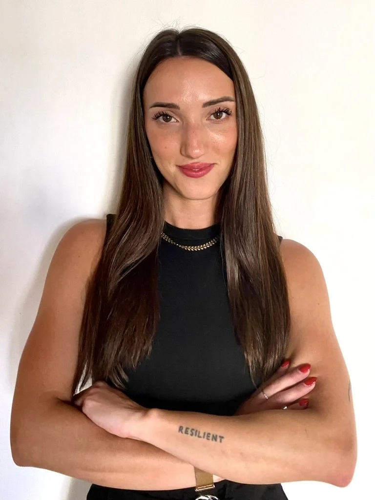 Elopement planner with long brown hair, wearing a black sleeveless top, gold necklace, and red nail polish, standing against a plain white wall with her arms crossed, displaying a tattoo that reads 'RESILIENT' on her left forearm.