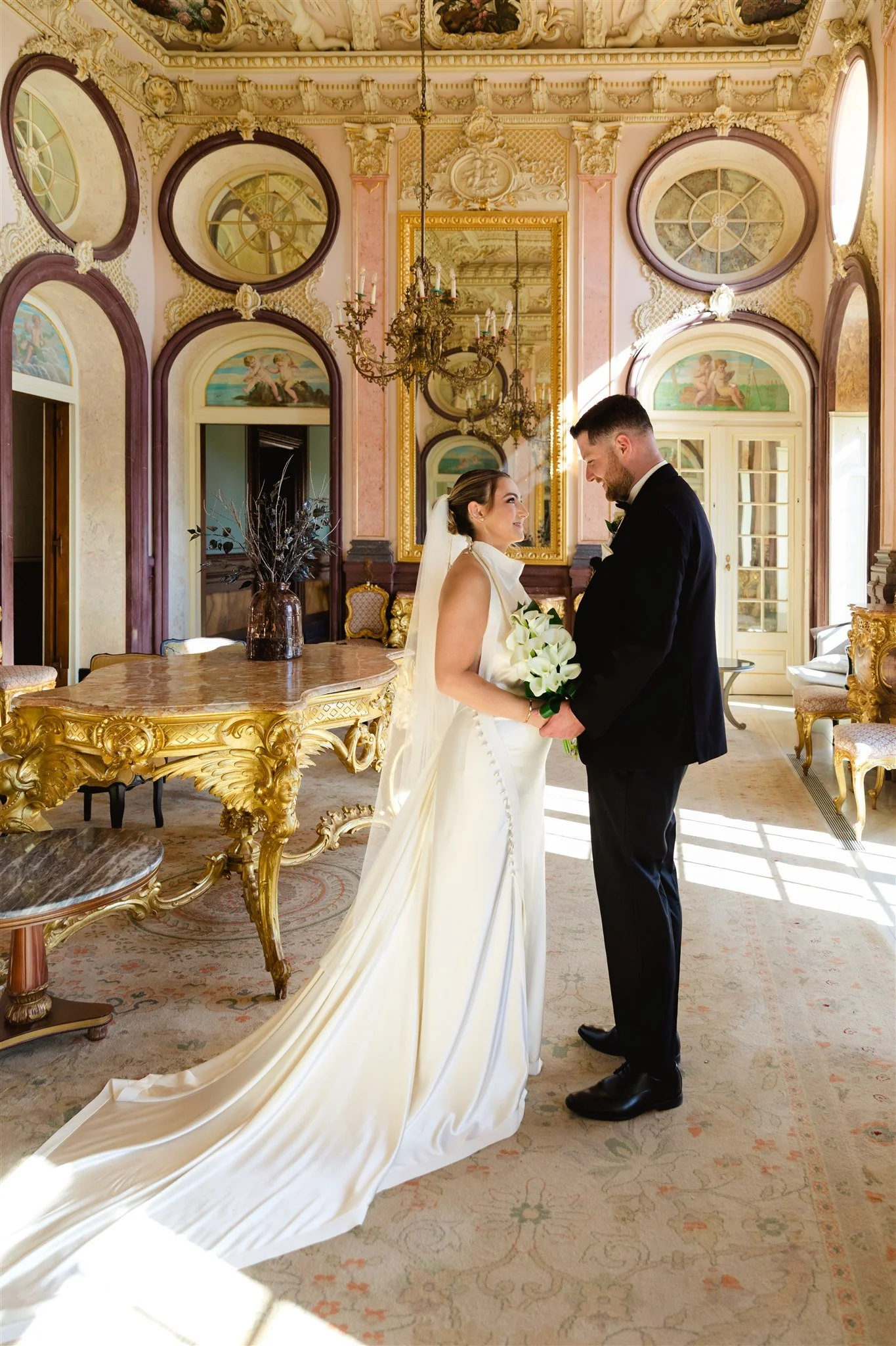 A bride and groom standing close together indoors, gazing at each other, in a luxurious, ornate room with chandeliers, large mirrors, and decorative painting details. The bride is holding a bouquet of white flowers, and the groom is dressed in a blac