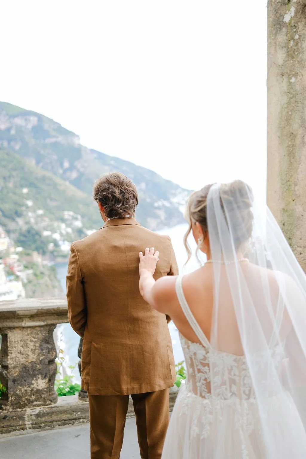 Bride touching groom's shoulder outdoors with mountains and water in the background.