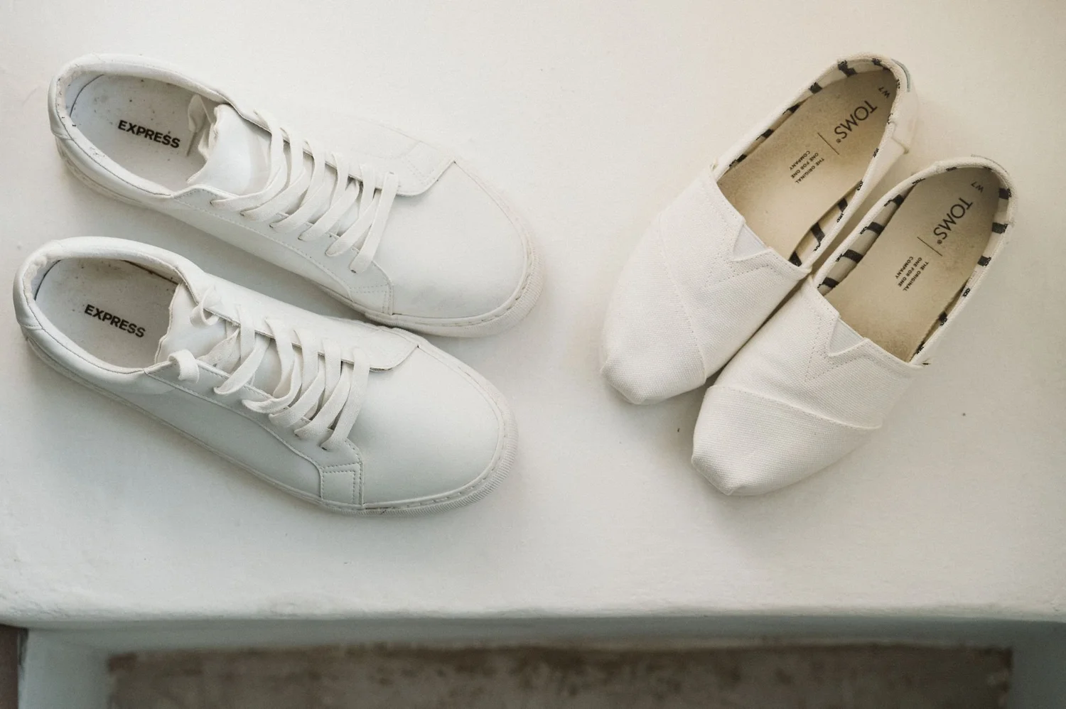 A pair of white sneakers with white laces and another pair of white slip-on shoes with black and white striped lining. The sneakers are on a white surface, seen from above.