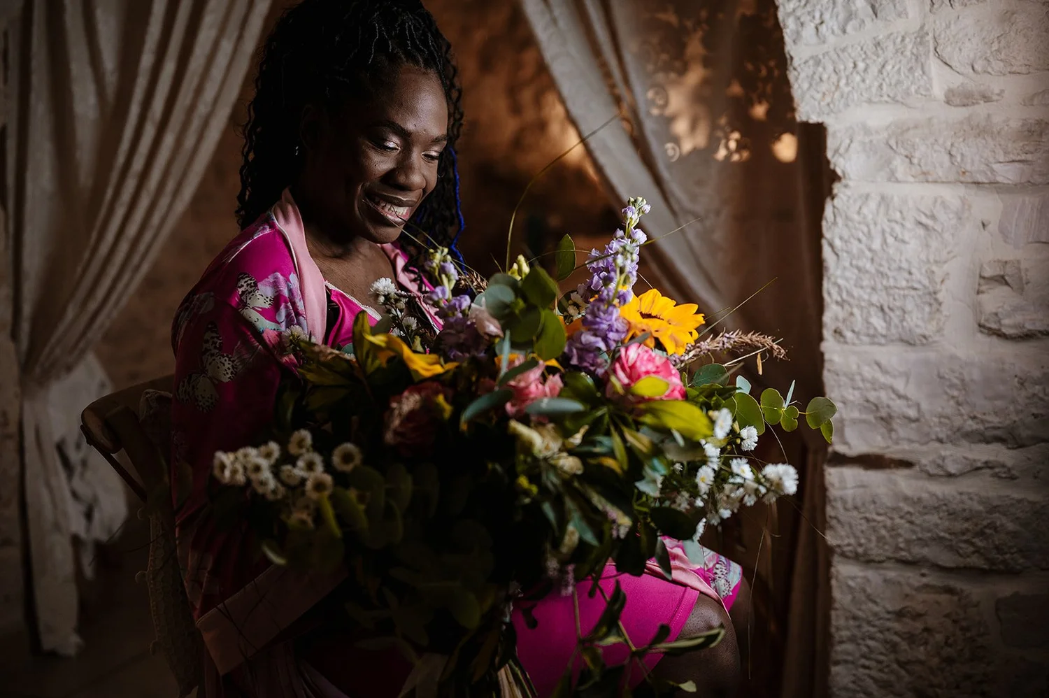 A woman in a pink dress sitting in a rustic room, holding a large bouquet of colorful flowers and smiling.