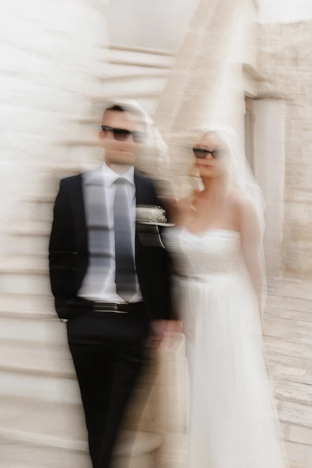 Blurred photo of a man in a suit and a woman in a wedding dress standing against a beige stone wall, both wearing sunglasses.