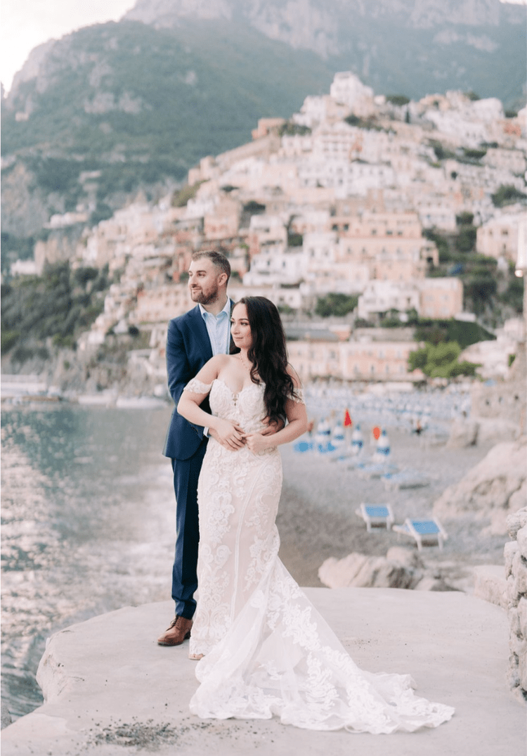 A newlywed couple in wedding attire poses by the water with a hillside town in the background, featuring pastel buildings and boats on the beach.