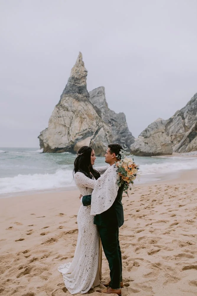 A couple dressed in wedding attire stands on a sandy beach, embracing and looking into each other's eyes, with a large rock formation and ocean waves in the background.