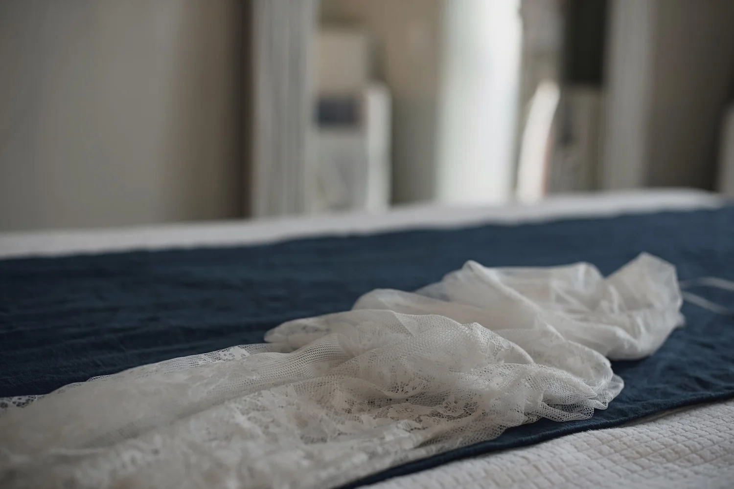 Close-up of white lace fabric on bed with dark blue striped blanket underneath, blurred background of a window with blinds.