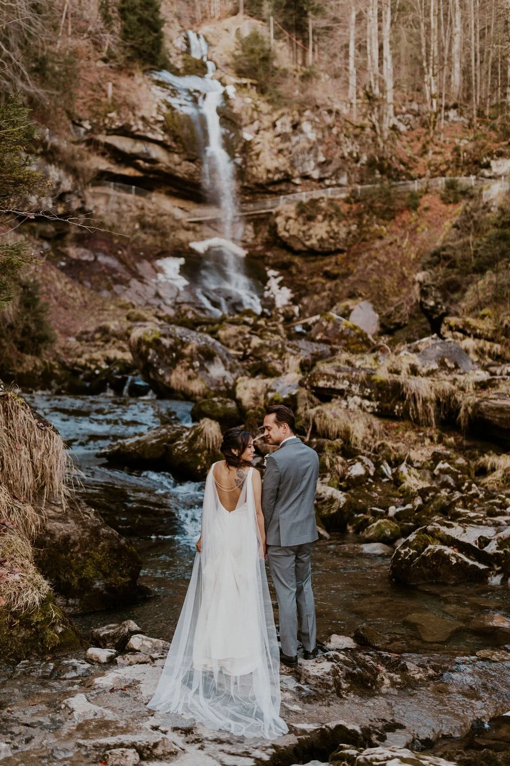 A bride and groom in wedding attire standing by a creek with rocks, with a waterfall and a forest backdrop in the background.