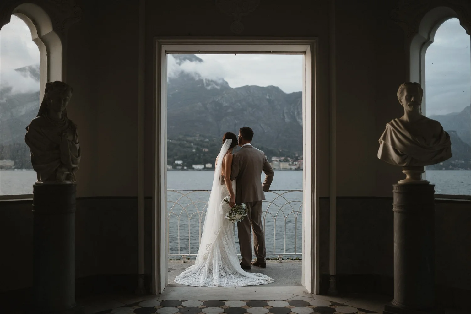 A bride and groom stand close together on a balcony overlooking a body of water and mountains, framed by a large window with ornate architectural details. Two bust sculptures are on either side of the window.