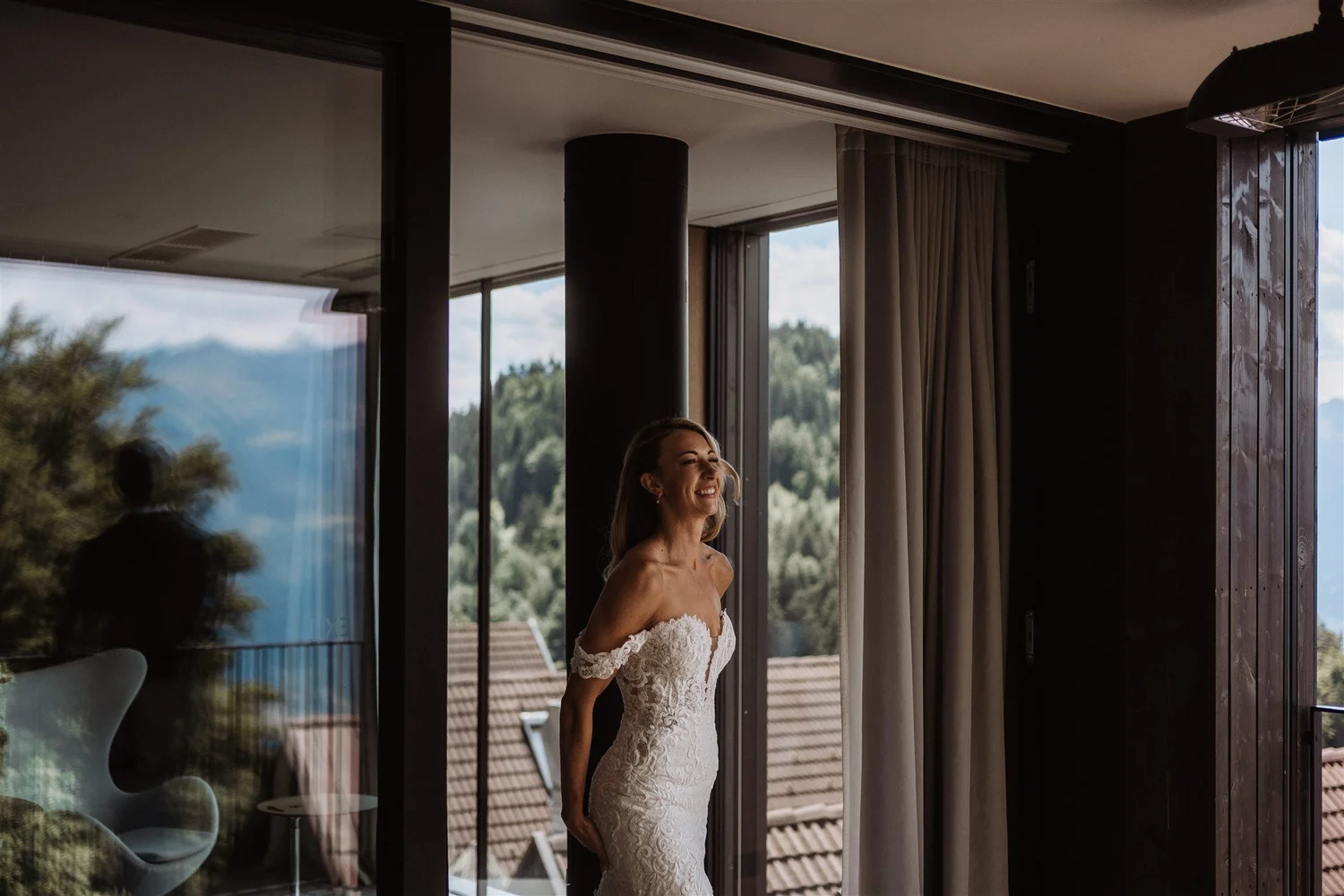 A woman in a white lace off-shoulder wedding dress standing near large glass windows with a scenic mountain landscape in the background.