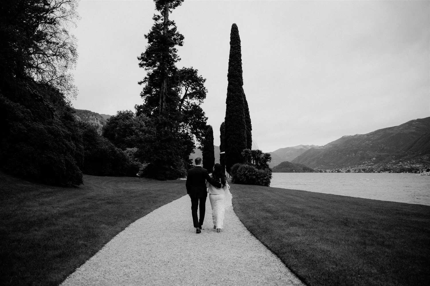 A black-and-white photo of a couple walking arm in arm along a gravel path in a scenic outdoor area near a lake. The background includes tall trees and mountain landscape.