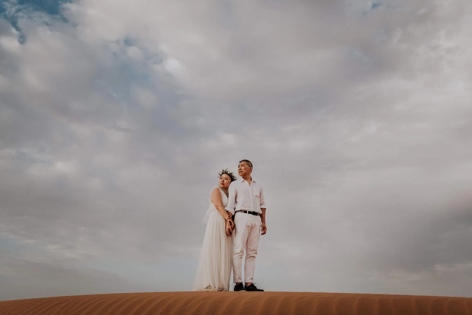 A couple stands close together in a desert landscape with a cloudy sky above. The woman wears a white dress and a floral crown, and the man wears a white shirt and light-colored pants.
