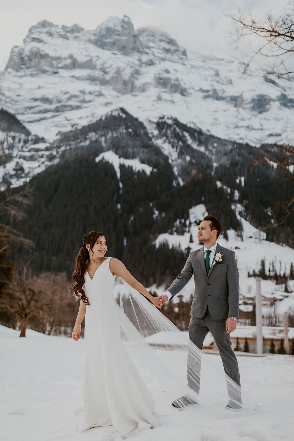 A bride and groom holding hands in a snowy landscape with a large snow-covered mountain in the background.
