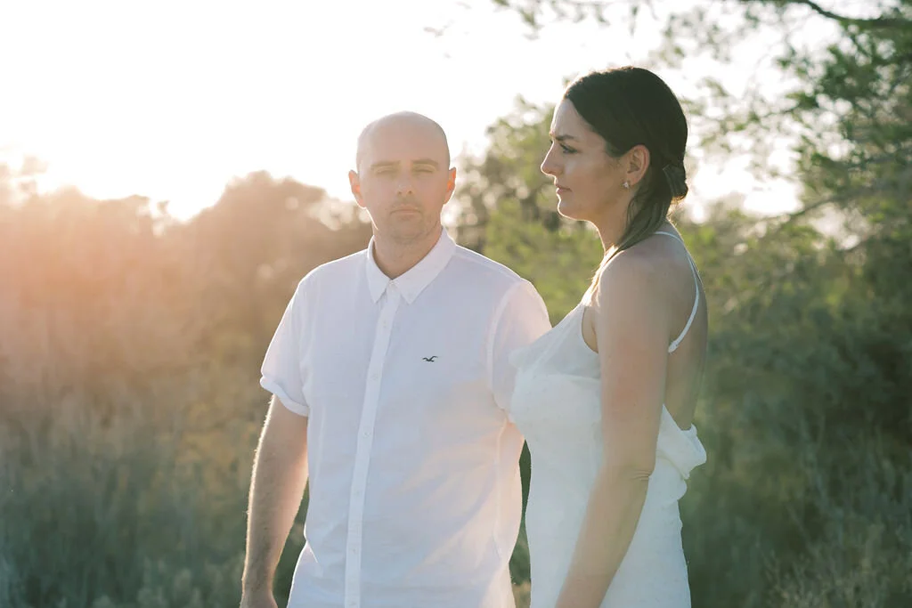 A man and a woman standing outdoors in a grassy area, with trees in the background, during sunset.