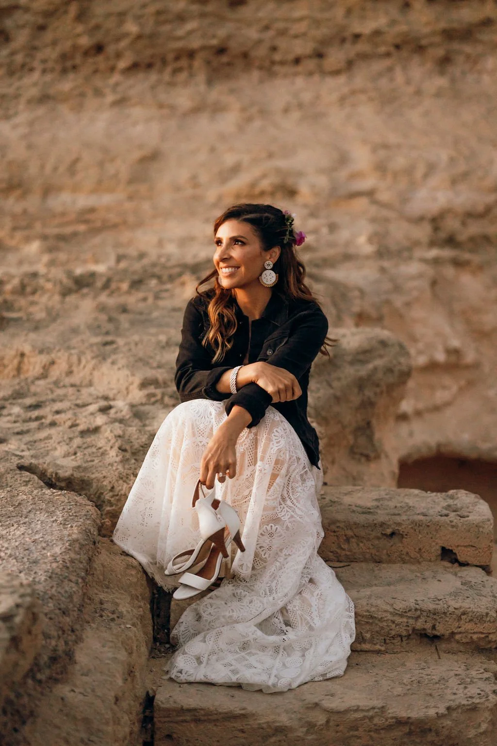 A woman in a white lace dress, black jacket, and large earrings sitting on stone steps outdoors, holding white high heels, smiling and looking to her left with flowers in her hair.