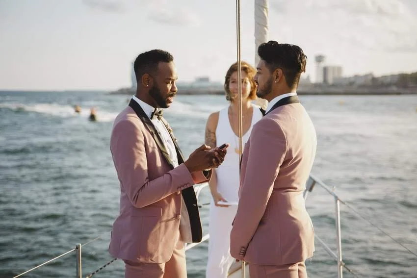 Two men in pink suits exchange vows on a boat, with a woman in a white dress standing behind them during a wedding ceremony by the water.