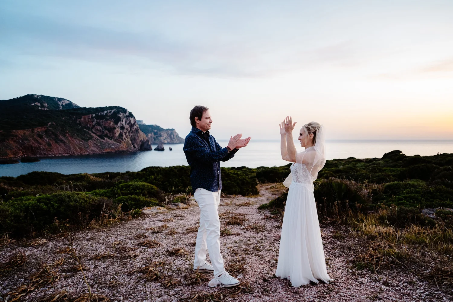 A couple having a wedding ceremony outdoors near the ocean during sunset, with mountains and greenery in the background. The groom claps as the bride smiles, raising her hand.