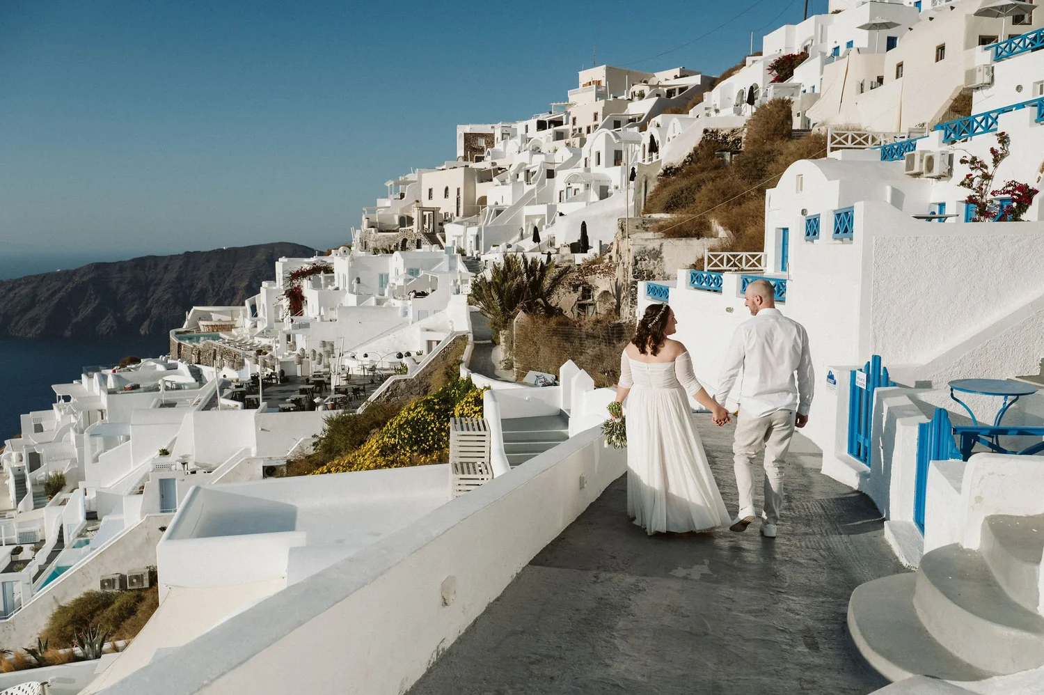 A bride and groom walking hand in hand along a white pathway on a hillside in Santorini, Greece, with white buildings and blue accents in the background.