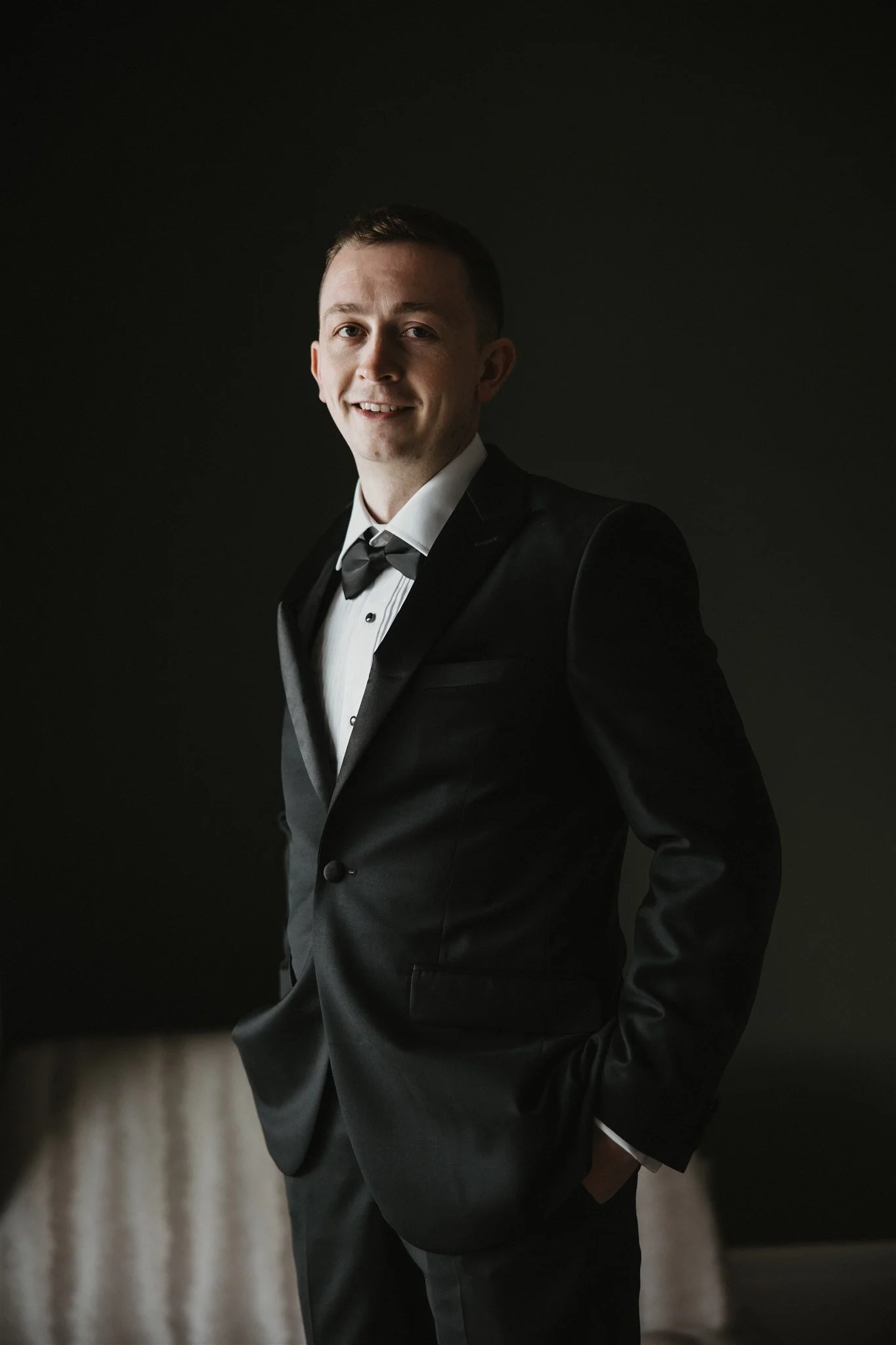 A young man in a tuxedo, smiling, standing with one hand in his pocket against a dark backdrop.