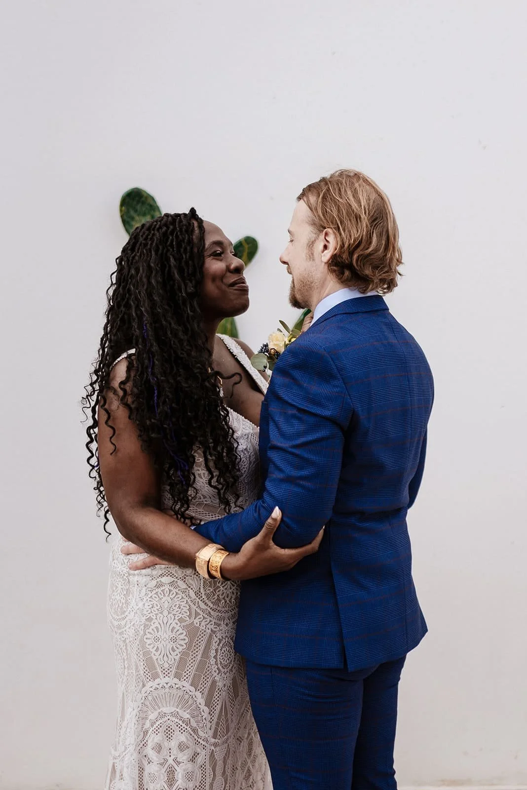 A couple, a woman with dark curly hair and a man with red hair, sharing a dance in wedding attire against a plain white wall, with a small green plant in the background.