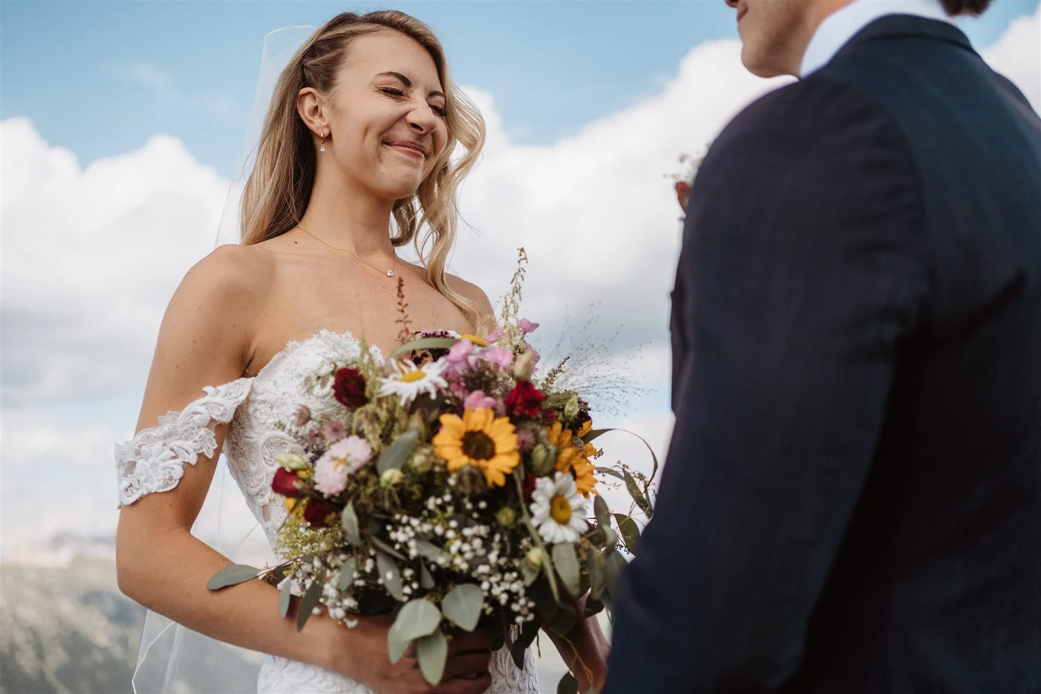 A bride is smiling and holding a bouquet of colorful flowers, standing outdoors with a blue sky and clouds in the background, as she faces a man dressed in a dark suit.