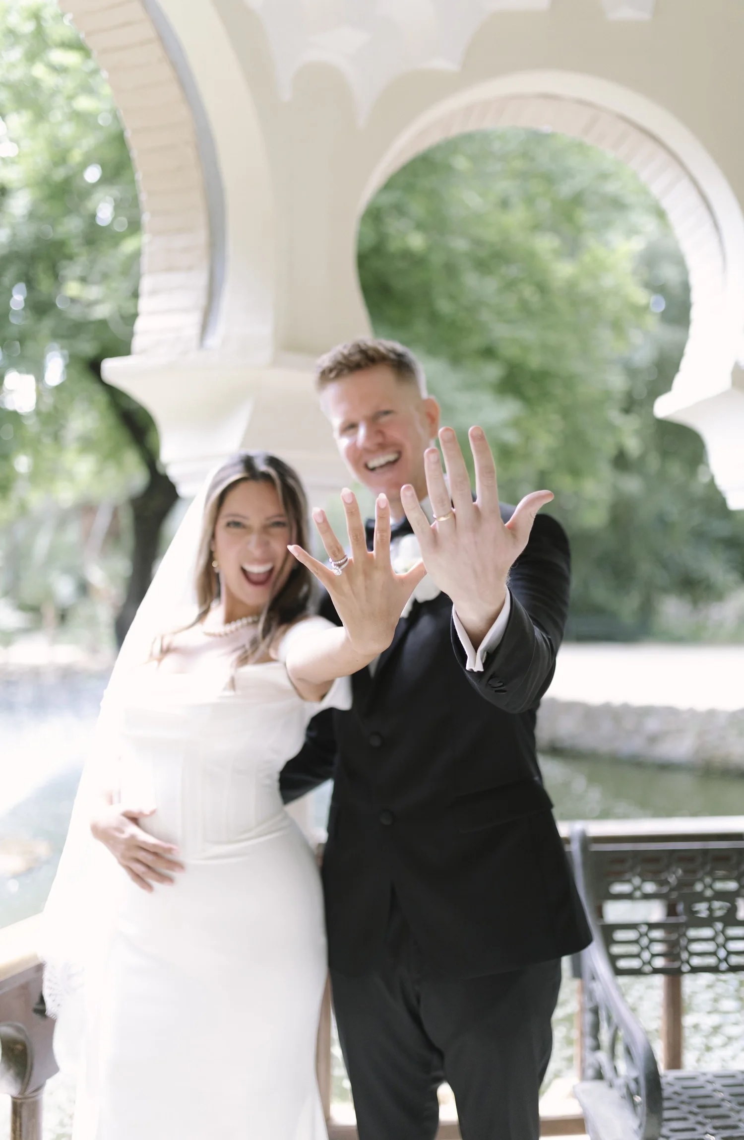 A newlywed couple is smiling and showing their wedding rings while holding hands near a park waterway with trees and a bridge in the background.