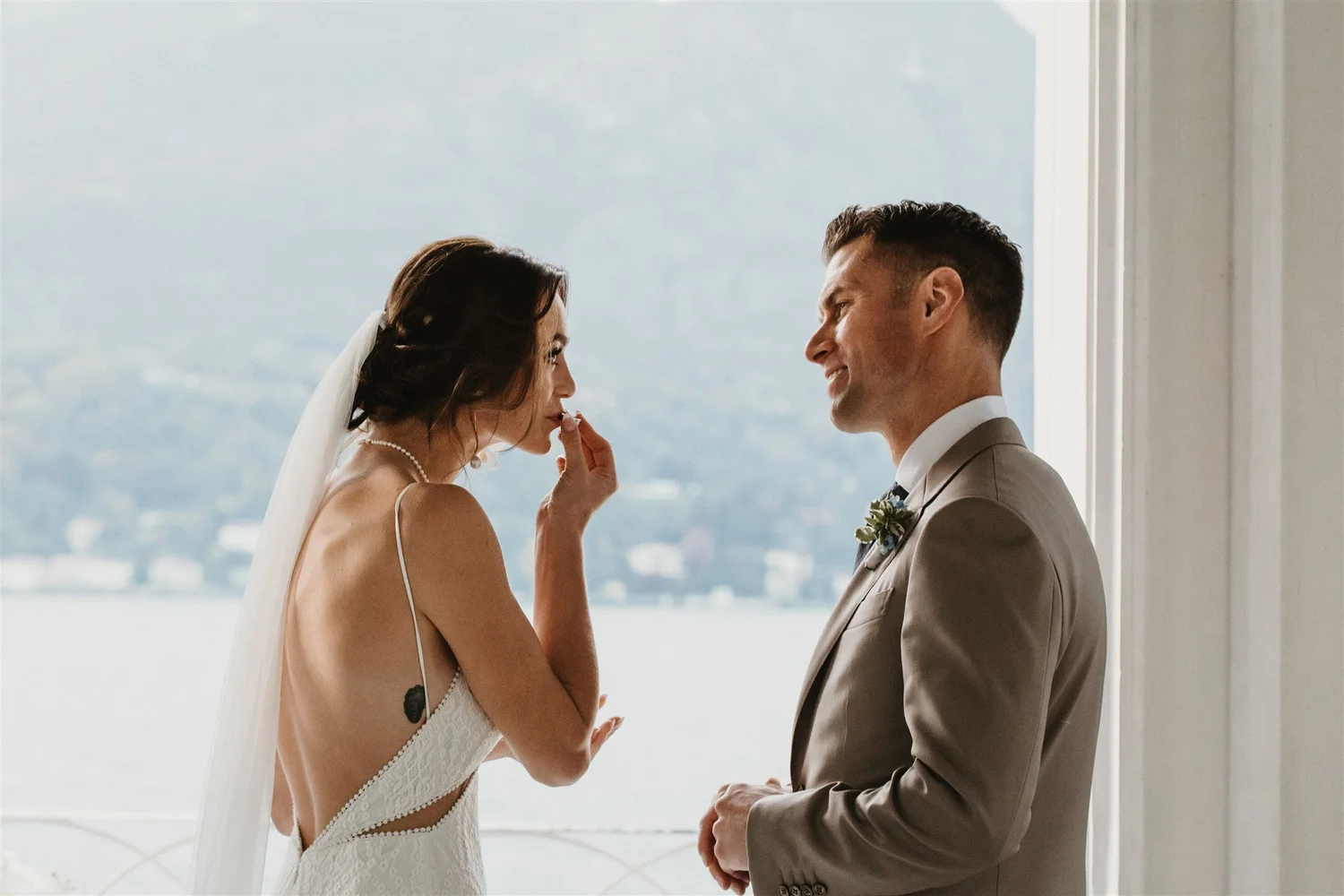 A bride with dark brown hair in a veil and a white lace dress is touching her lips as she looks into the eyes of a groom with short, dark hair who is smiling and wearing a beige suit with a white shirt and a boutonniere. They are standing indoors nea