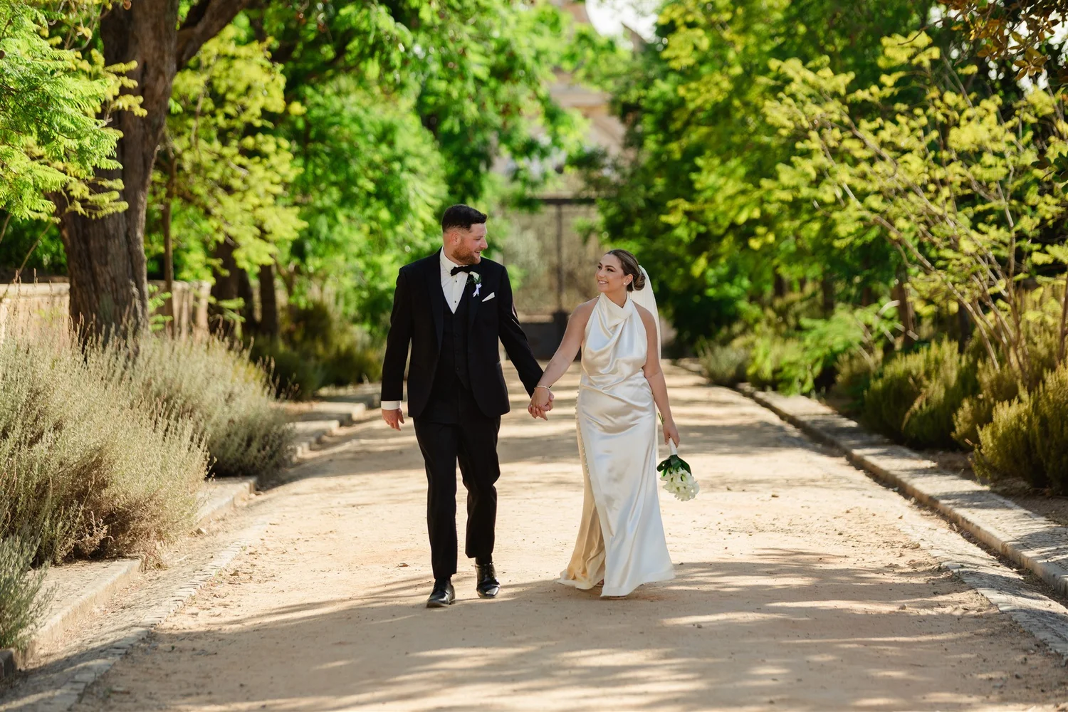 A newlywed couple walking hand in hand on a tree-lined path, smiling at each other, surrounded by green foliage.