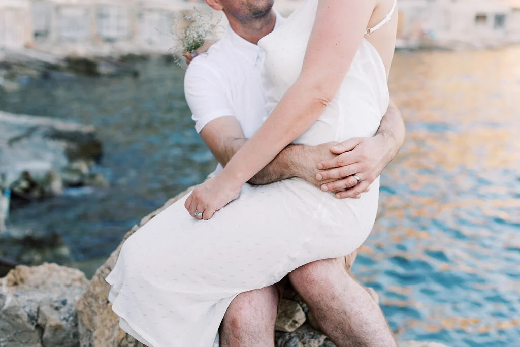 A man is holding a woman in his arms near a body of water, with rocks and rippling water in the background. The woman is wearing a white dress, and the man has a ring on his finger.