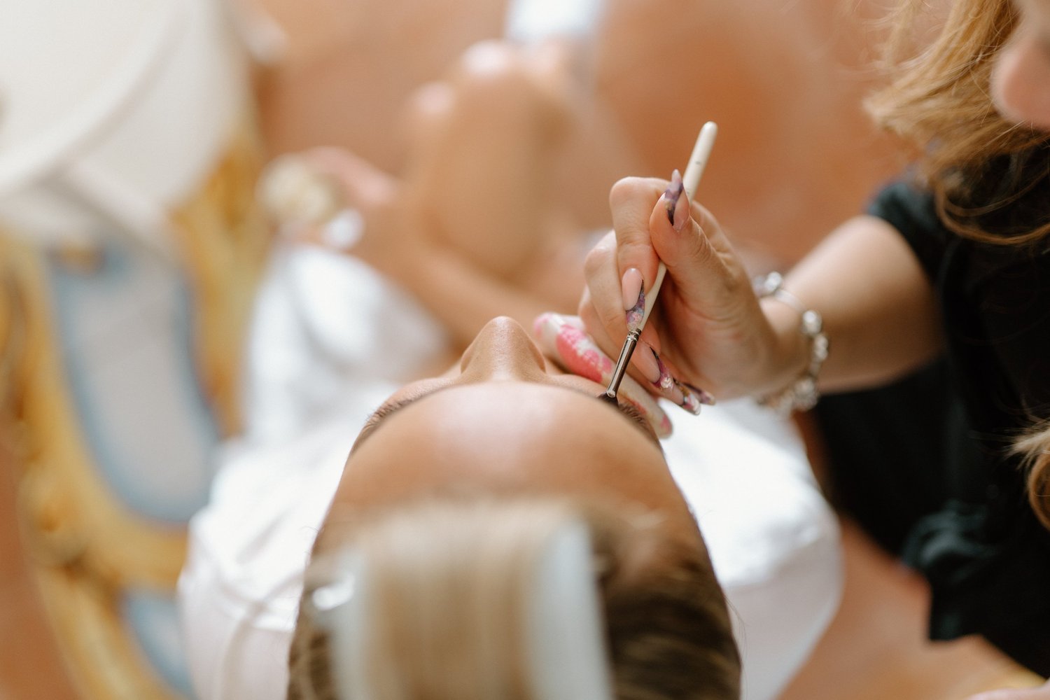 A person with long, dark hair performing a nail art design on another person's hand, using a fine brush, in a close-up shot.