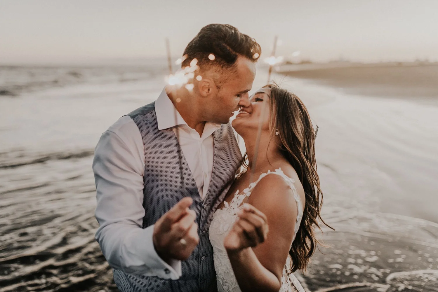 A couple dancing closely on a beach during sunset, with waves in the background and light reflecting on the water.