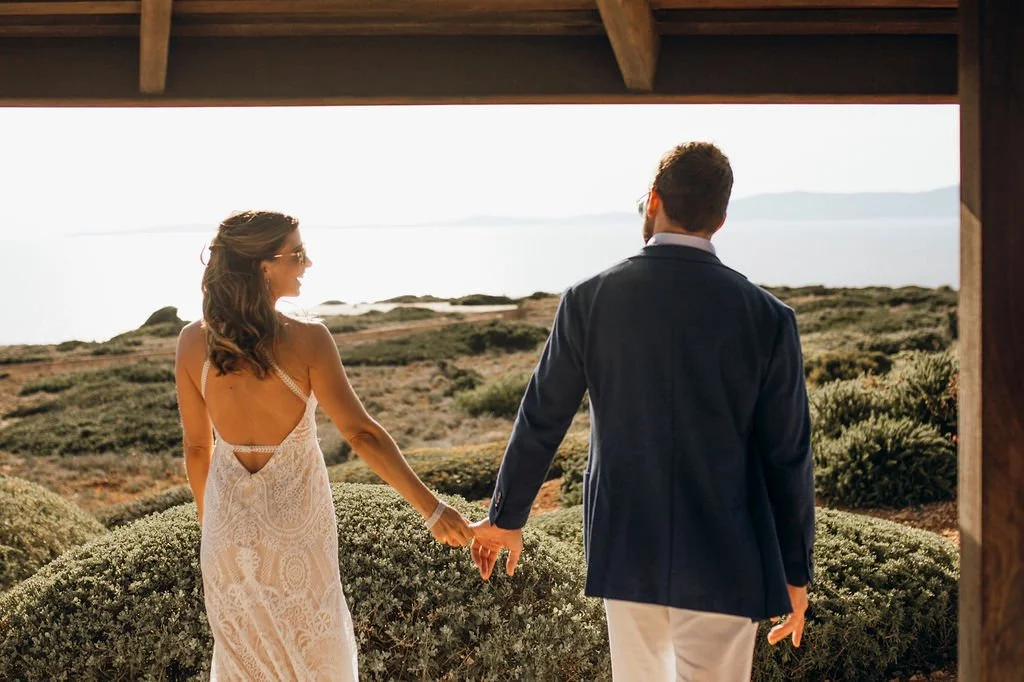 A couple dressed in formal wedding attire holding hands and walking outside near the ocean with a scenic landscape and cloudy sky in the background.