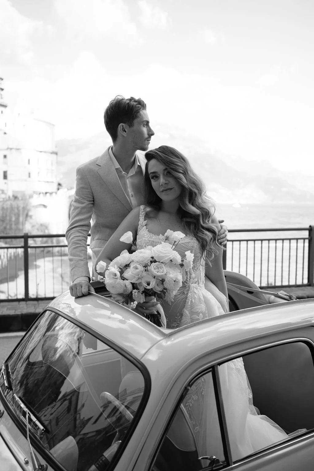 Black and white photo of a couple, bride holding a large bouquet of roses, standing in a vintage car with a scenic outdoor background featuring water and mountains.