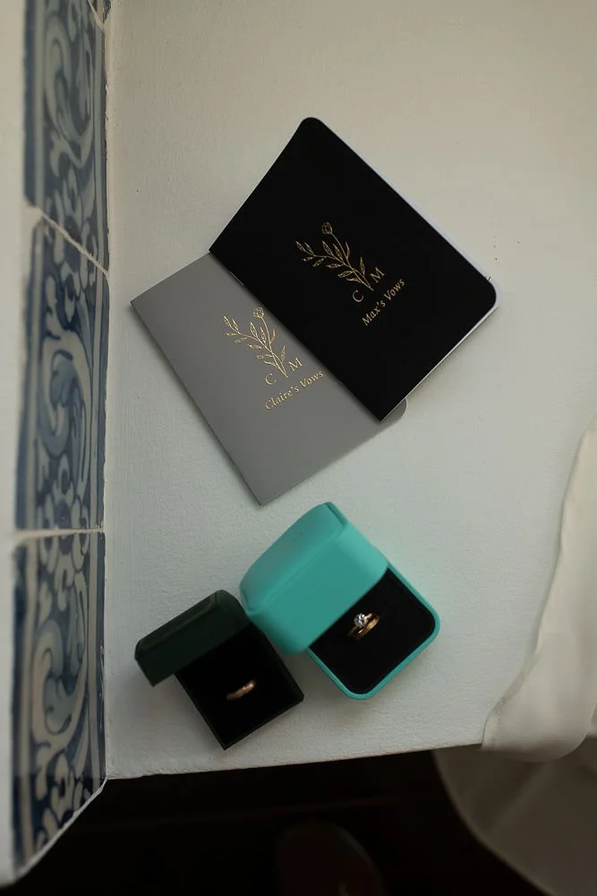 Two jewelry boxes, one black and one turquoise, with rings inside. Two greeting cards with gold lettering and botanical designs, one black and one gray, reading 'C & M Max's Vows' and 'C & M Claire's Vows.'