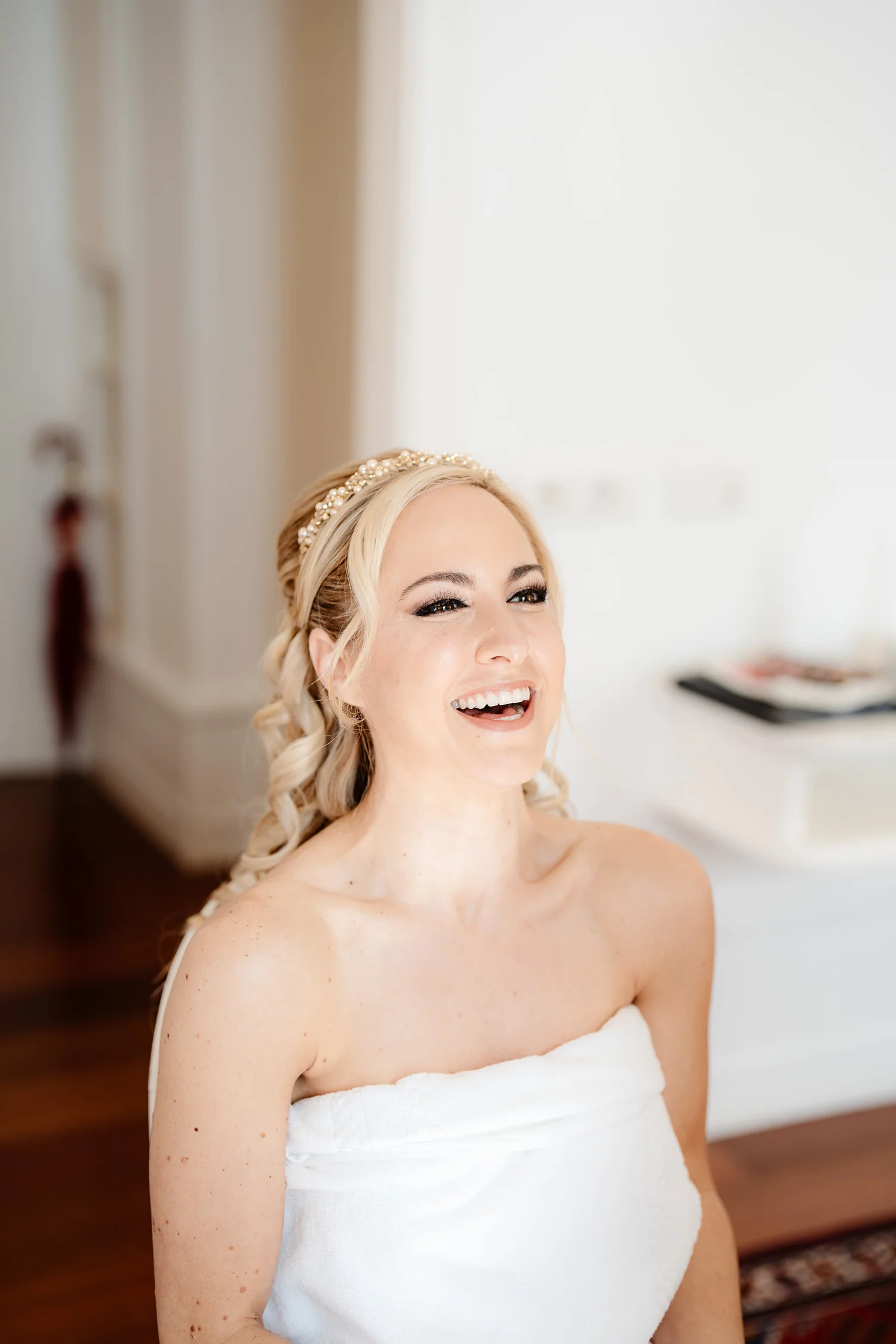 A woman with blonde hair styled in loose curls, wearing a pearl headband, smiling and wrapped in a white towel.
