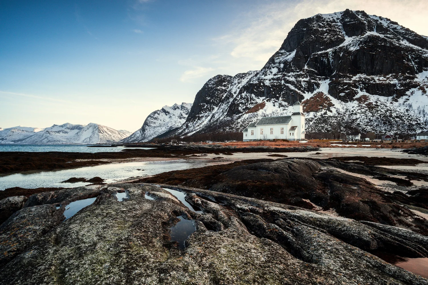 Snow-capped mountains with a rocky shoreline and a white church near the water, under a partly cloudy sky.
