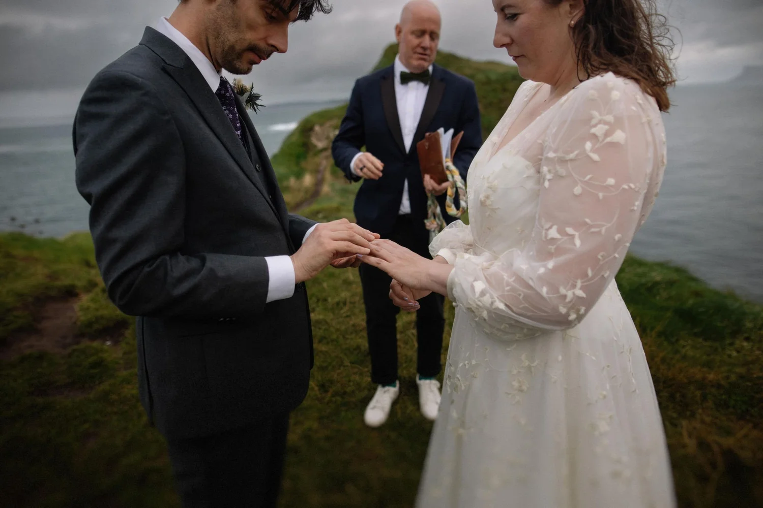 A man and woman exchanging wedding vows outdoors near a body of water with a hilly landscape in the background, accompanied by an officiant holding a book.