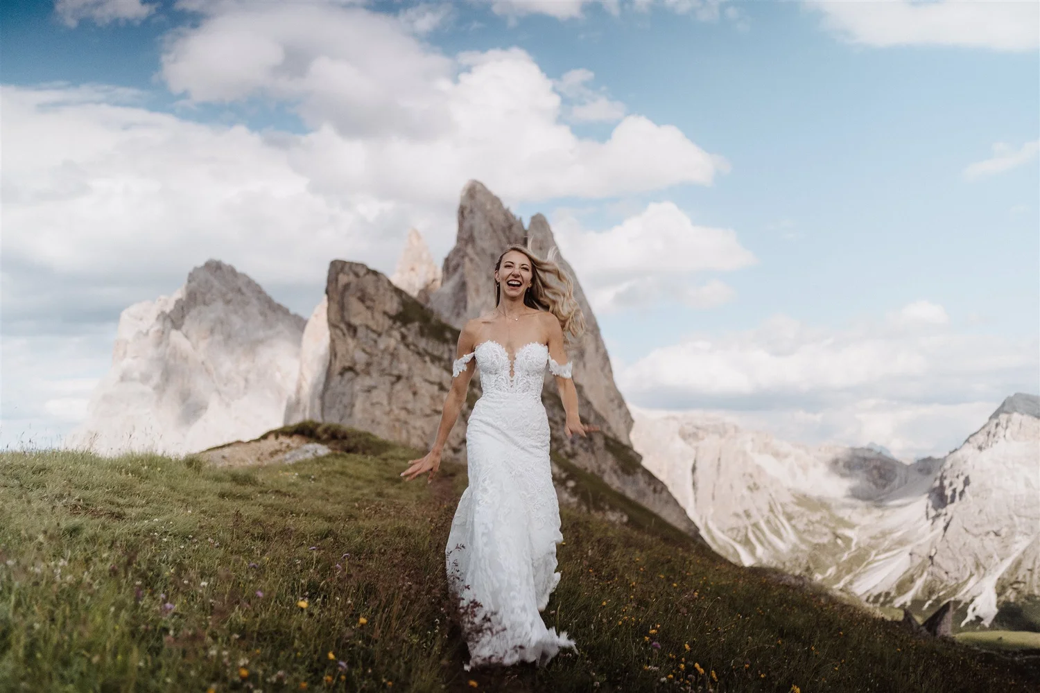 A woman in a white wedding dress standing on a grassy area with mountains and clouds in the background, smiling and looking joyful.