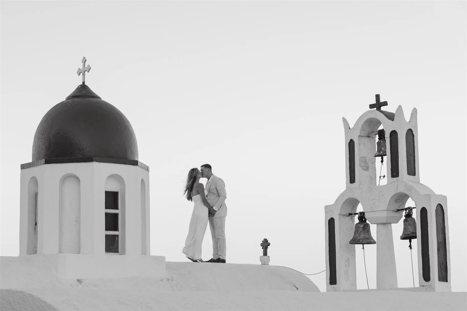 A couple stands close together on a white rooftop between two small white church buildings with domed roofs and crosses, in a black and white photo.