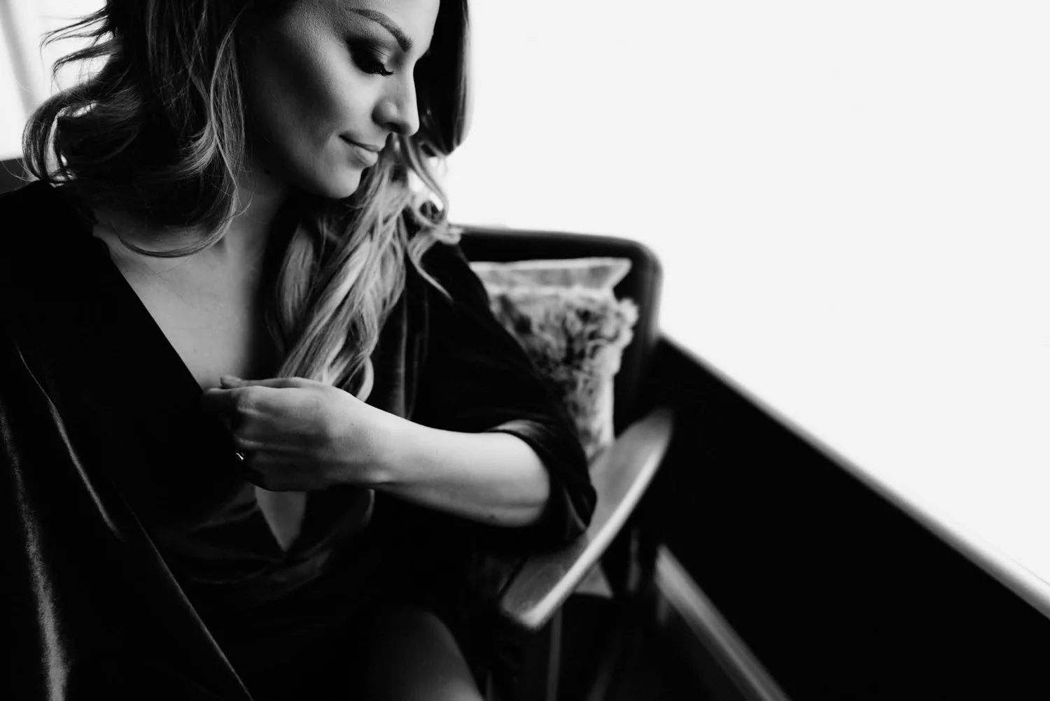 A black-and-white photo of a woman with long hair, sitting and smiling with her eyes closed, holding a cup, in a cozy indoor setting.