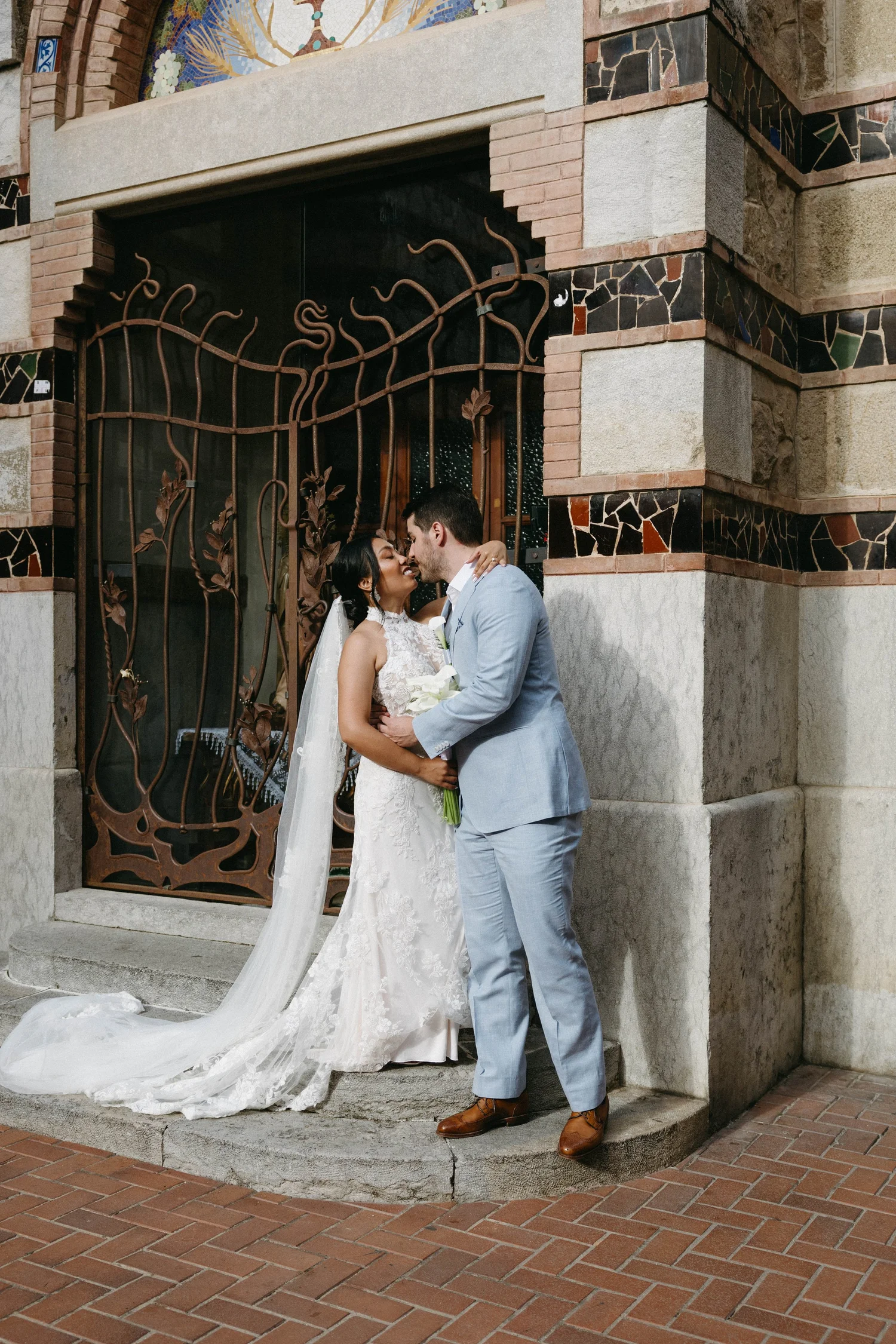 A bride and groom kissing outside a building with a decorative iron gate and mosaic artwork. The bride is in a white lace wedding dress with a long train, and the groom is in a light gray suit with brown shoes.