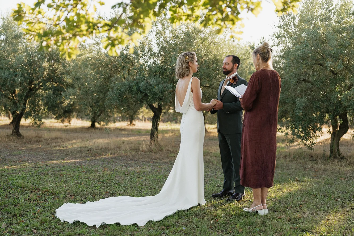 A couple getting married outdoors during daytime, with a person officiating and trees in the background.