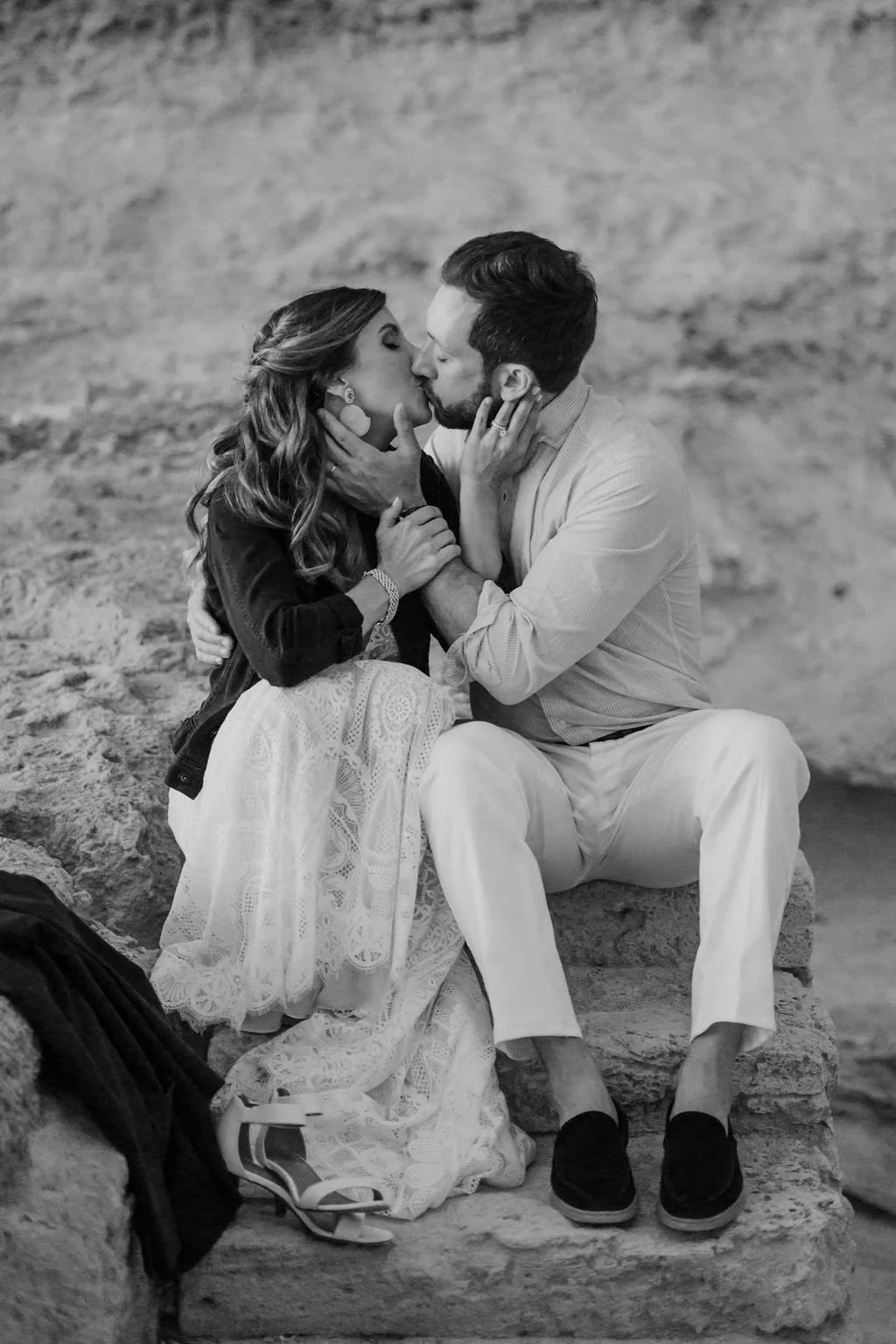 A black and white photo of a couple kissing, sitting on a stone ledge outdoors with a blurred rocky background. The woman has long wavy hair, wearing earrings, a dark top, and a lacy skirt. The man has short dark hair, wearing a light shirt and white pants. Their hands are touching, and a pair of shoes and a bag are on the ground beside them.