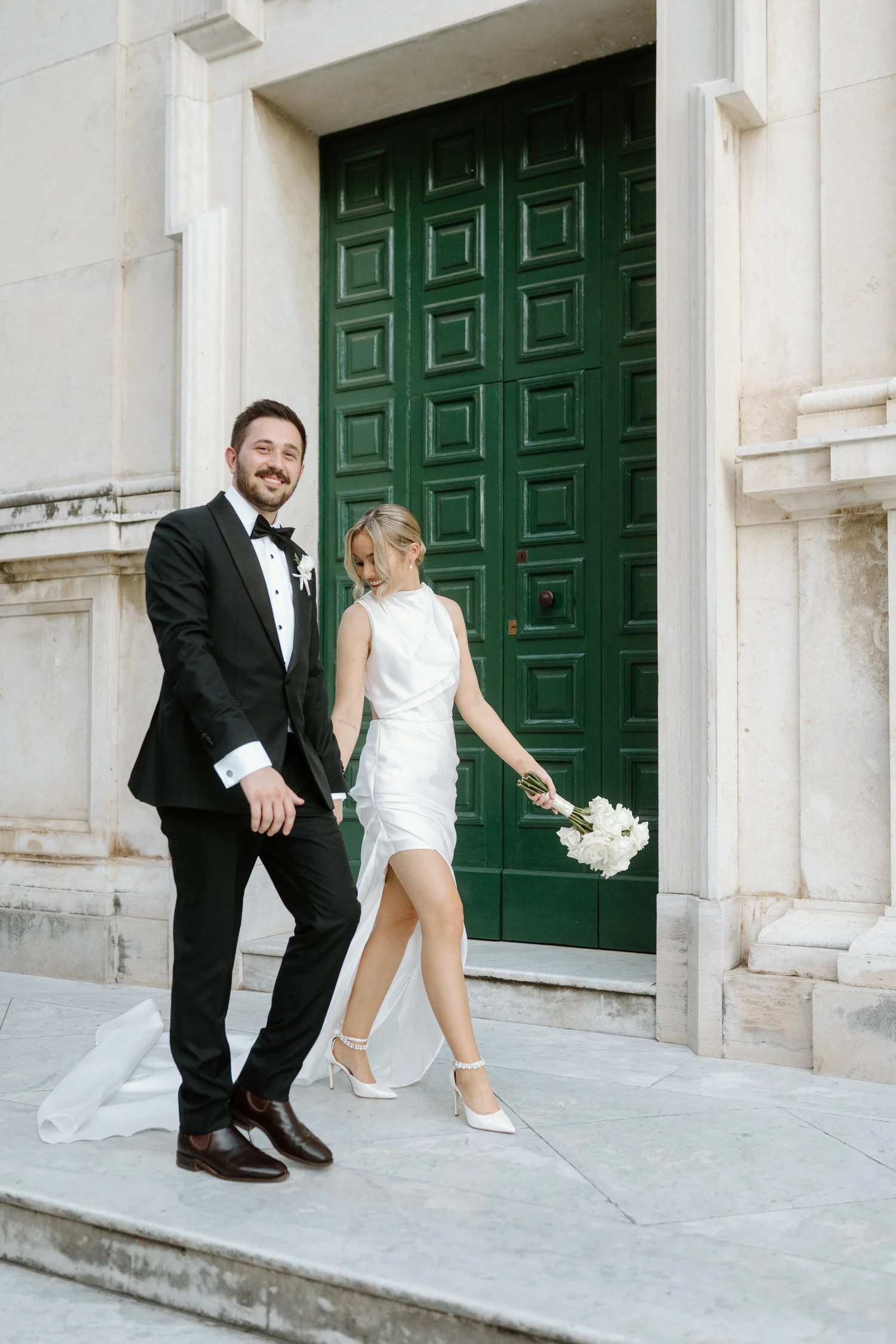 A newlywed couple walking down the steps of a building with large green doors. The groom is wearing a black tuxedo and bow tie, and the bride is dressed in a white wedding gown holding a bouquet of white flowers.