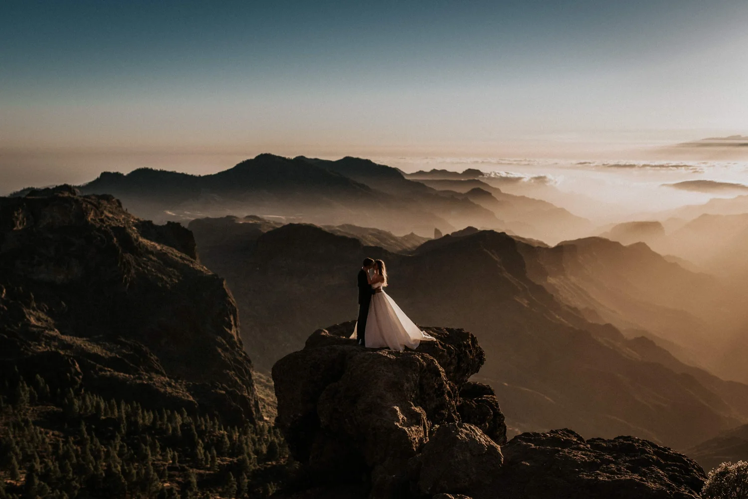 A bride and groom standing on a rocky cliff, embracing each other with a mountain range and sunset in the background.