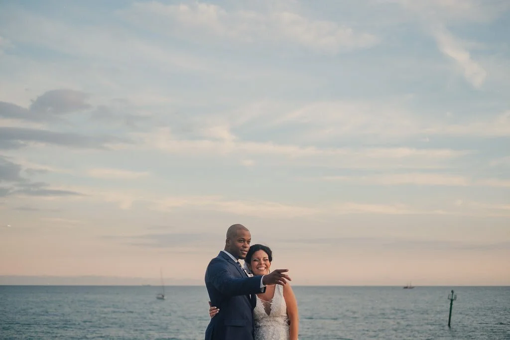 A couple dressed in wedding attire standing together on a beach at sunset with the ocean and sailboats in the background.