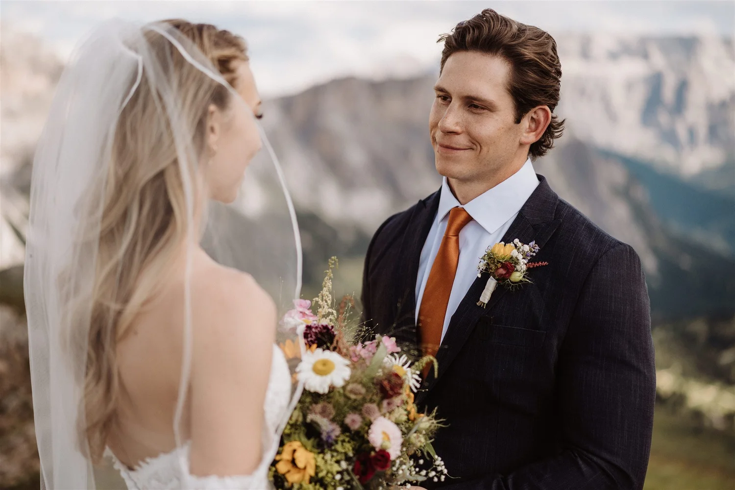 A bride and groom at their wedding, facing each other outdoors, with mountains in the background. The bride is wearing a white wedding dress and veil, holding a colorful bouquet, while the groom is dressed in a dark suit with a white shirt and an ora