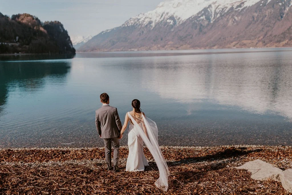 A bride and groom standing hand in hand on a pebbled beach, looking out at a tranquil lake surrounded by mountains with snow caps.