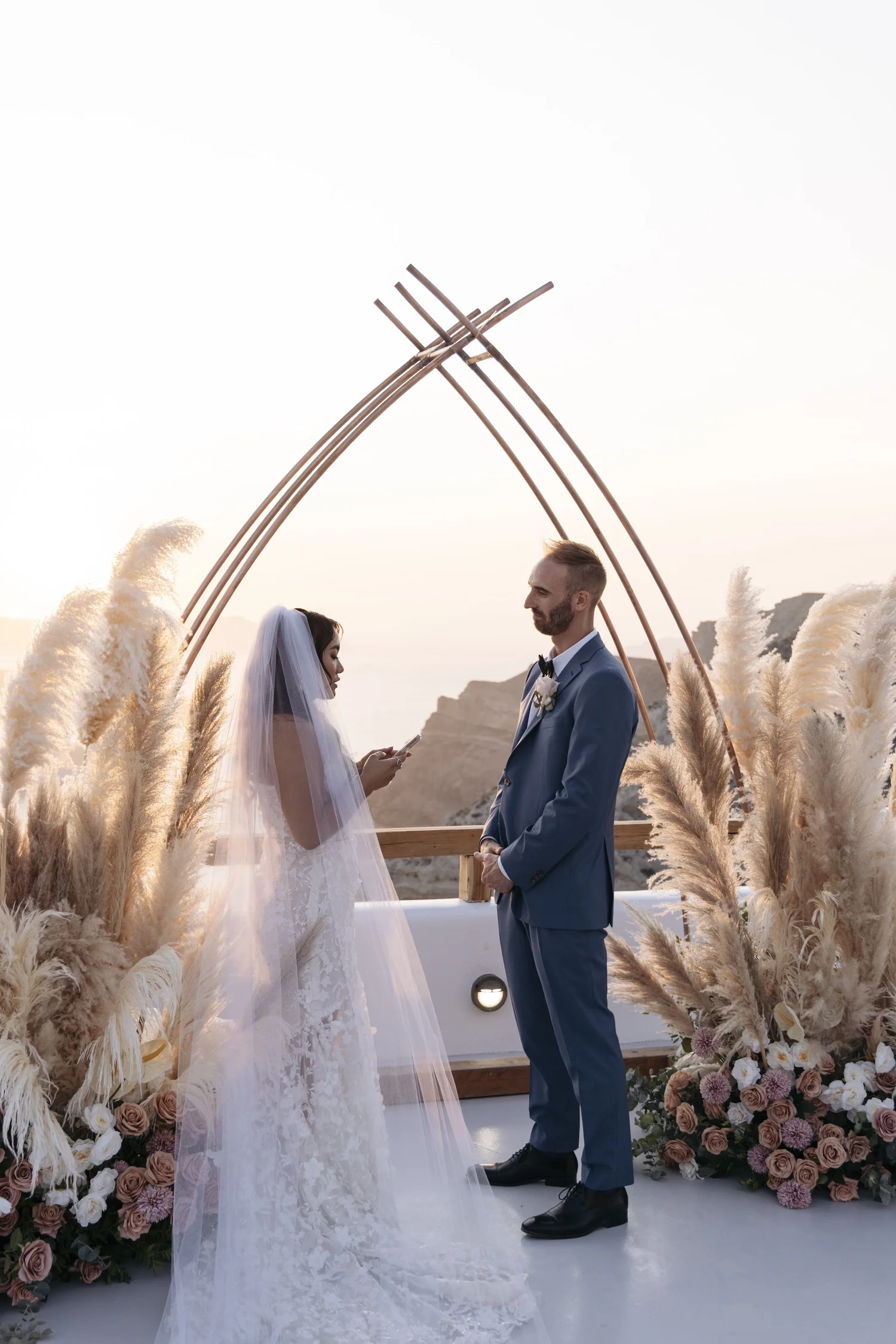 A bride and groom standing face to face during their outdoor wedding ceremony at sunset, surrounded by pampas grass and floral arrangements.
