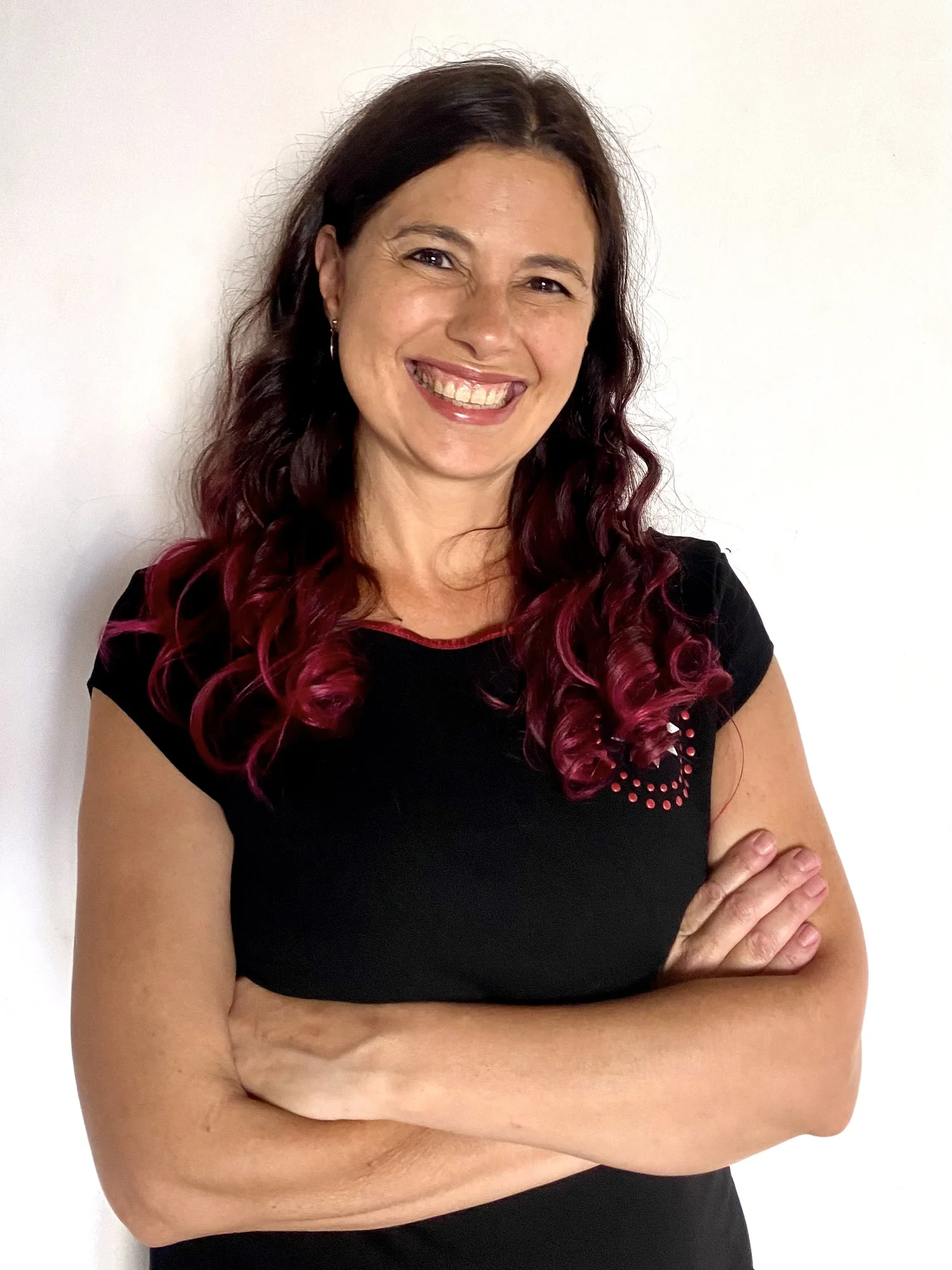 Elopement planner with dark brown hair and pinkish highlights wears a black top with small pink and white design and is crossing her arms in front of a plain white wall, smiling at the camera.