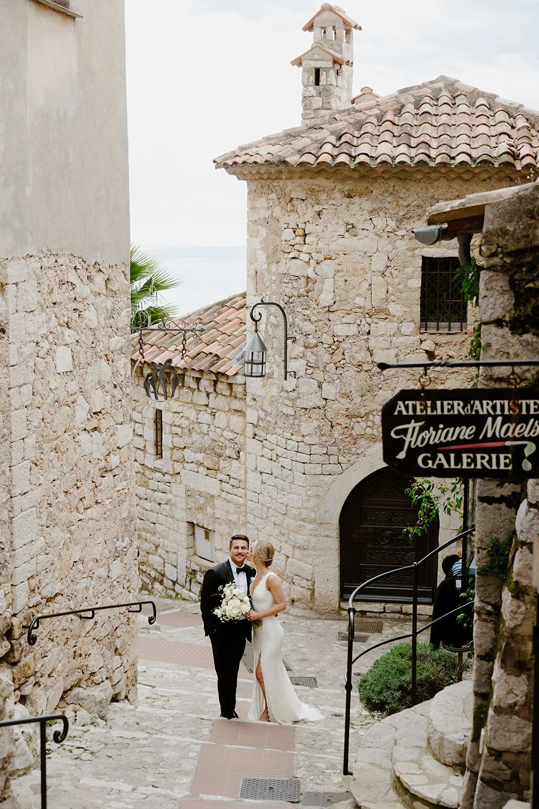 A bride and groom standing on a cobblestone street in front of a stone archway during daytime. The groom is in a black tuxedo with a bow tie, holding a bouquet of white flowers. The bride is in a white lace wedding gown, gazing at the groom. The setting is a historic European-style town with old stone buildings and a sign for an art gallery.