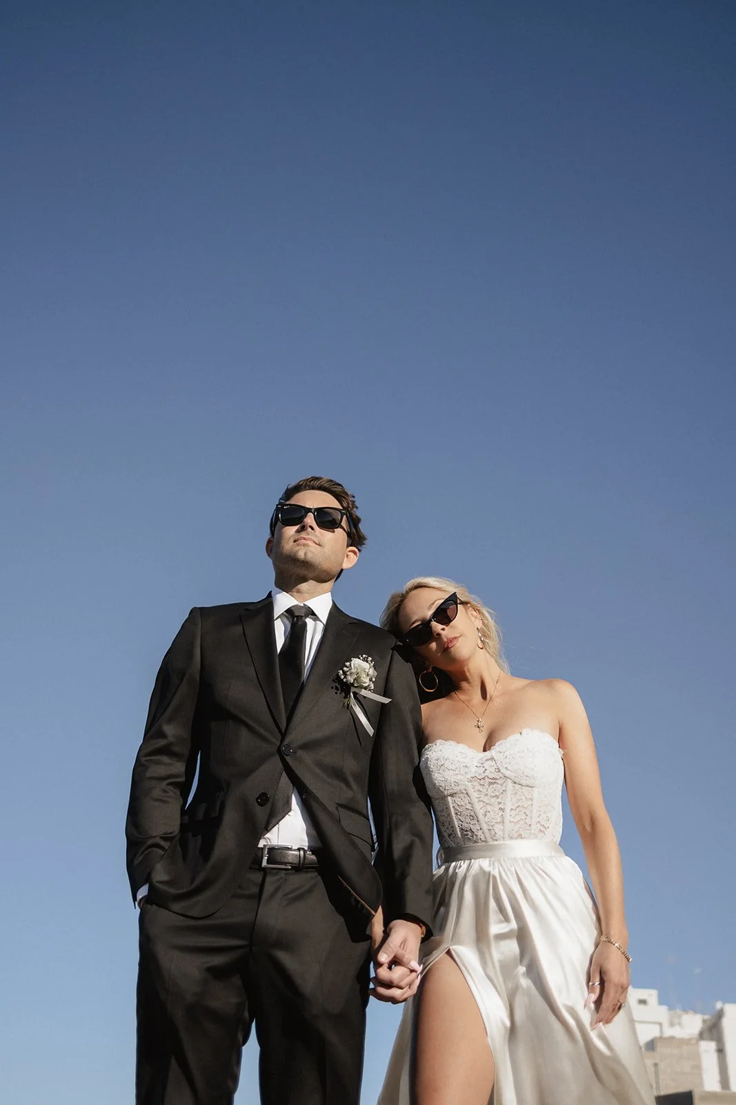 A man and woman dressed in wedding attire, holding hands, standing outdoors against a clear blue sky. The man is in a black suit and tie, wearing sunglasses, and the woman is in a strapless white wedding dress with a slit, also wearing sunglasses.